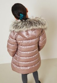 Chaqueta tipo plumón en rosa claro con rayas horizontales, que cuenta con una capucha de pelo sintético desmontable y un tejido suave y brillante. Leggings gris suave son visibles.
