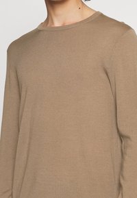 Hellbrauner langärmliger Pullover aus weichem Stoff mit einem gerippten runden Halsausschnitt und subtilen Nähten an den Ärmeln.