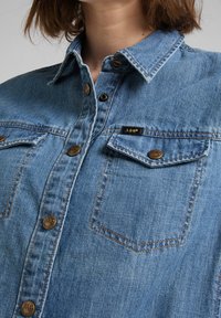 Denimskjorta i ljusblått med klassisk krage, kopparfärgade knappar, två bröstfickor och en diskret logolapp på vänster ficka.