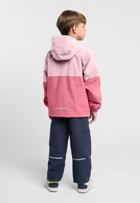 Giacca impermeabile rosa e rosa chiaro con cappuccio, polsini elastici e dettagli riflettenti, abbinata a pantaloni blu e sneakers bianche.