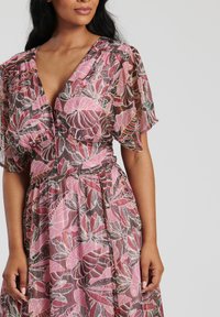 Blommig klänning i rosa och brunt med korta, luftiga ärmar. Tyget har ett livfullt bladmönster och en figursydd midja. Knappdetalj framtill.
