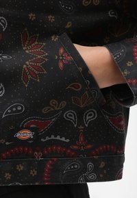 Svart tygjacka som har ett färgglatt paisley-mönster i röd, guld och grått, med en vävd etikett vid fållen.