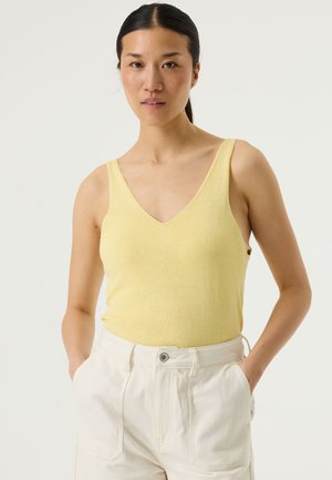 Geel mouwloos topje met geribbelde textuur en V-hals. Gecombineerd met hooggetailleerde crèmekleurige broek met steekzakken aan de voorkant en een knoopsluiting.