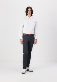Chemise blanche à manches longues avec deux poches poitrine, associée à un pantalon bleu marine et des baskets grises avec des accents noirs.