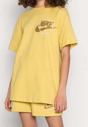 Persoon draagt een geel Nike T-shirt en bijpassende short met een bruin Nike-logo en witte cirkelvormige tekst op de borst en het been.