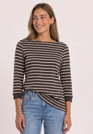 Frau lächelt seitlich, trägt ein braun-beige gestreiftes Langarmshirt und hellblaue Jeans vor einem einfarbigen hellen Hintergrund.