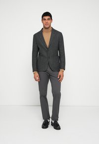 Blazer cinzento com lapelas entalhadas, camisola de gola alta castanha clara, calças cinzentas e sapatos pretos, posicionado contra um fundo branco.