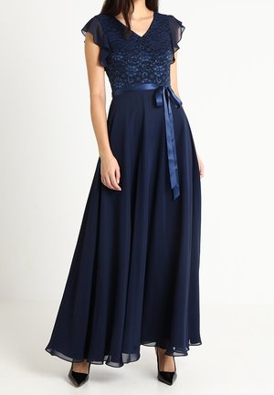 Donna che indossa un abito lungo blu navy con corpetto in pizzo, maniche corte svolazzanti e una cintura in raso in vita, abbinato a tacchi neri.