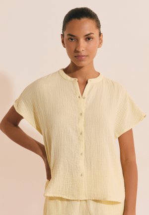 Kobieta o ciemnych włosach w kremowej, teksturowanej koszuli z krótkim rękawem i guzikami, pozująca na jasnobeżowym tle.