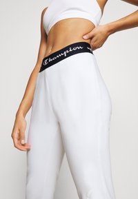 Białe spodnie dresowe z czarnym elastycznym pasem w talii z logo "Champion". Gładka tekstura i kieszenie po bokach. Modelka pozuje z ręką na biodrze.