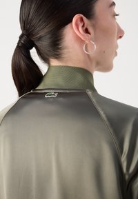 Olivenfarget, lett jakke med høy krage, glatt tekstur, som har en liten brodert krokodillelogo på baksiden.