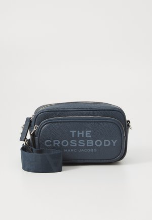 Marine farget læder crossbody taske med tekstureret overflade, der har to lynlåslommer og trykt branding på forsiden. Justerbar rem inkluderet.