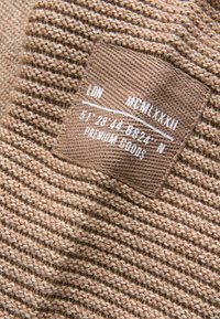Beige gestrickter Stoff mit strukturierten Rillen, versehen mit einem braunen Etikett mit weißem Text, der Koordinaten und "PREMIUM GOODS" angibt.