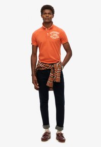 Pomarańczowa koszulka polo z białym logo, ciemne dżinsy, kraciasta koszula przewiązana w pasie oraz burgundowe buty z beżowymi sznurówkami.