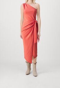 Robe corail à une épaule avec tissu drapé, nouée à la taille, avec une fente sur le côté. Portée avec des bottes de cheville beiges, texture lisse.
