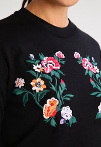 Musta neulepaita, jossa värikäs kukkaiskirjailu punaisena, oranssina, valkoisena ja vaaleanpunaisena. Resoroitu pyöreä kaulus ja pehmeä neulospinta.