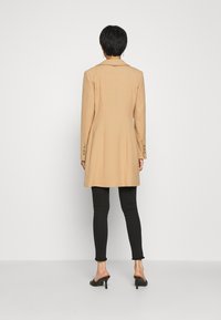 Cappotto lungo beige con un design sartoriale, dotato di colletto, chiusura a singolo bottone e polsini con bottone, indossato su leggings neri e scarpe con tacco.