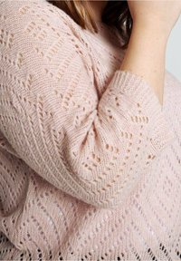 Pull tricoté rose clair avec des motifs texturés en mailles ouvertes, manches trois-quarts, et une apparence douce et duveteuse.