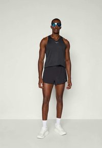Svart atletisk linne och shorts, lättviktigt tyg, med blå reflexglasögon och vita sneakers, står framför en neutral bakgrund.
