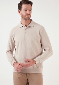 Beige hosszú ujjú pólóing, texturált anyagból készült, klasszikus gallérral és három gombos záródással, bordázott mandzsettákkal és aljjal.