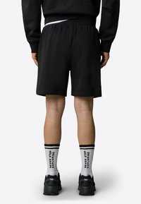 Los pantalones cortos deportivos negros tienen una cinturilla elástica. Se combinan con calcetines blancos que tienen rayas y texto en negro, y zapatillas negras.