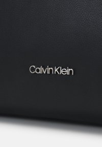 Svart texturerad läderhandväska med silverfärgad "Calvin Klein"-logotyp. Har en elegant design och en slät yta. Klassisk och mångsidig form.