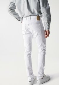 Białe dżinsy o slim fit, z tylnymi kieszeniami i skórzaną metką w kolorze brązowym na pasku. Noszone z białymi sneakersami i jasnoszarym t-shirtem.