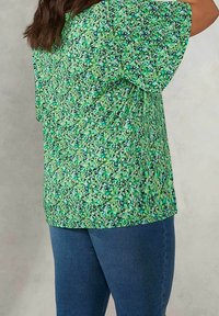 Krátký rukáv zelená květinová blůza s různými odstíny a vzory, doplněná modrými džíny. Tkanina vypadá lehce a texturovaně.