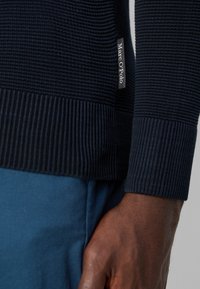 Marineblauer, gerippter Pullover mit strukturiertem Strick, taillierten Bündchen und einem Etikett mit der Aufschrift "Marc O'Polo". Kombiniert mit hellblauen Hosen für einen Kontrast.