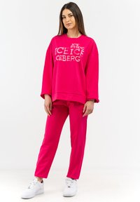 Felpa rosa con grafica "ICE ICE ICEBERG", abbinata a pantaloni rosa. Entrambi hanno una vestibilità rilassata. Il modello indossa sneakers bianche.