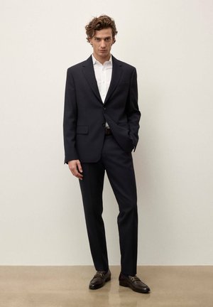 Egy férfi áll a fal mellett, sötét tengerészkék öltönyt visel, fehér inggel és barna bőr mokaszinokat fém csatos díszítéssel.