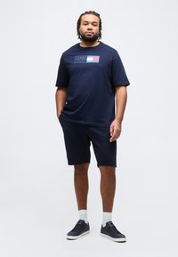 Άνδρας με κοτσιδωτά μαλλιά, που φοράει μπλε ναυτικό μπλουζάκι Tommy Hilfiger, αντίστοιχα σορτς, λευκές κάλτσες και μπλε αθλητικά παπούτσια, στέκεται μπροστά σε απλό φόντο.