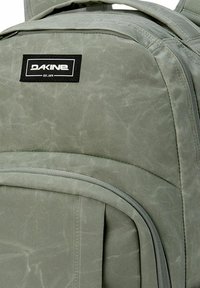 Dakine-reppu hennossa vihreässä kankaassa, jossa on tekstuuri, musta logo-patch ja vetoketjulliset osastot säilytystä varten.