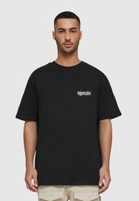 Sort bomulds T-shirt med korte ærmer, rund halsudskæring og hvidt tekstlogo på venstre bryst. Model bærer lysebeige bukser.