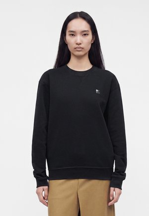 Frau mit langen schwarzen Haaren, die einen schwarzen Sweatshirt mit kleinem weißem Logo und beigen Hosen trägt, steht vor einem einfarbigen weißen Hintergrund.