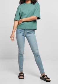 Luźna bawełniana koszulka w kolorze turkusowym z krótkim rękawem, zestawiona z jasnoniebieskimi dopasowanymi dżinsami i czarnymi sandałami z grubym, skrzyżowanym paskiem.