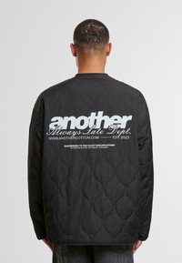 Black quilted jacket with a rounded neckline. Features prominent white graphic text on the back, and a smooth, shiny texture.