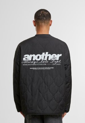 Black quilted jacket with a rounded neckline. Features prominent white graphic text on the back, and a smooth, shiny texture.