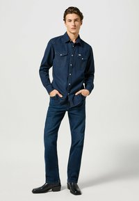 Mörkblå denimskjorta med tryckknappar, två framfickor och kontrasterande sömmar, kombinerad med mörka jeans och svarta ankelstövlar.