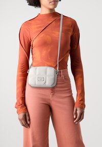 Borsa a tracolla in pelle grigia con motivo trapuntato e logo in tonalità argento. Il modello indossa un top arancione a maniche lunghe e pantaloni rosa.