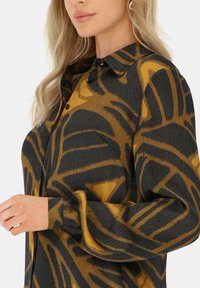 Bluzka z długim rękawem w kolorze czarnym i złotym, z fakturowanym materiałem o wyraźnym abstrakcyjnym wzorze, klasycznym kołnierzykiem i zapięciem na guziki z przodu.