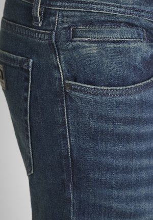Primo piano di una tasca di jeans in denim blu con cuciture dettagliate e rivetti sul lato destro.