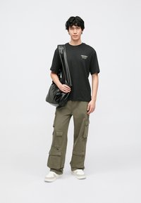 Black t-shirt with white text, olive cargo pants with side pockets, and beige sneakers. Model holds a black shoulder bag, standing on a plain background.