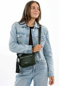 Piccola borsa a tracolla in pelle verde con superficie texturizzata, chiusura con zip e dettaglio frangi, abbinata a una giacca di jeans e jeans.
