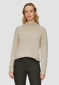 Beige Rollkragenpullover mit glatter Textur, langen Ärmeln und gerippten Bündchen, kombiniert mit dunkelgrüner Jeans, die vor einem schlichten Hintergrund posiert.