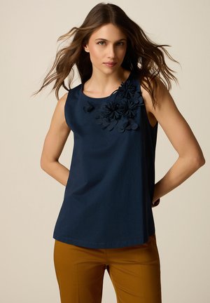Donna con capelli castano chiaro sciolti indossa un top senza maniche blu navy con applicazioni floreali e pantaloni color senape su uno sfondo beige uniforme.