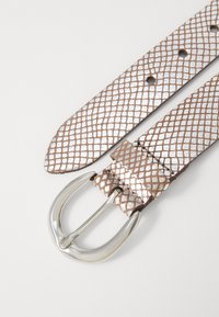 Metallisk tan lærbelte med diamantmønster, sølvspenne og flere hull for justering. Teksturert overflate gir økt visuell interesse.