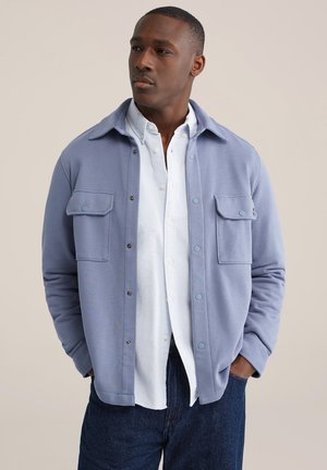 Lichtblauw jack met knopen, twee borstzakken, een kraag en zachte stof. Gedragen over een wit shirt en donkere spijkerbroek.