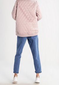 Lys rosa quiltet jakke med ribbet kant, parret med løse blå denimjeans og hvide sneakers, set fra ryggen mod en ensfarvet baggrund.
