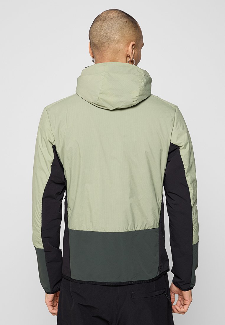 Homme avec la tête rasée portant une veste à capuche vert clair et noir, montré de dos sur un fond gris uni.